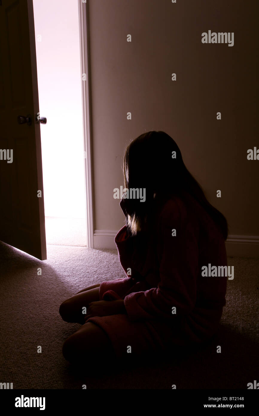Young girl sitting in a dark room, back view door open. Stock Photo