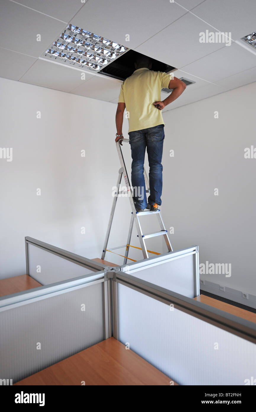 Man on top of step ladder assessing an electric problem in the false ceiling of an office room Stock Photo