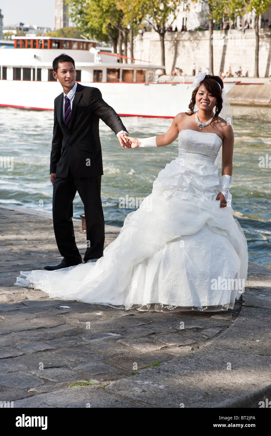smiling sun dappled Asian bride in strapless gown & handsome groom pose for portrait on Paris quai against blue waters of Seine Stock Photo