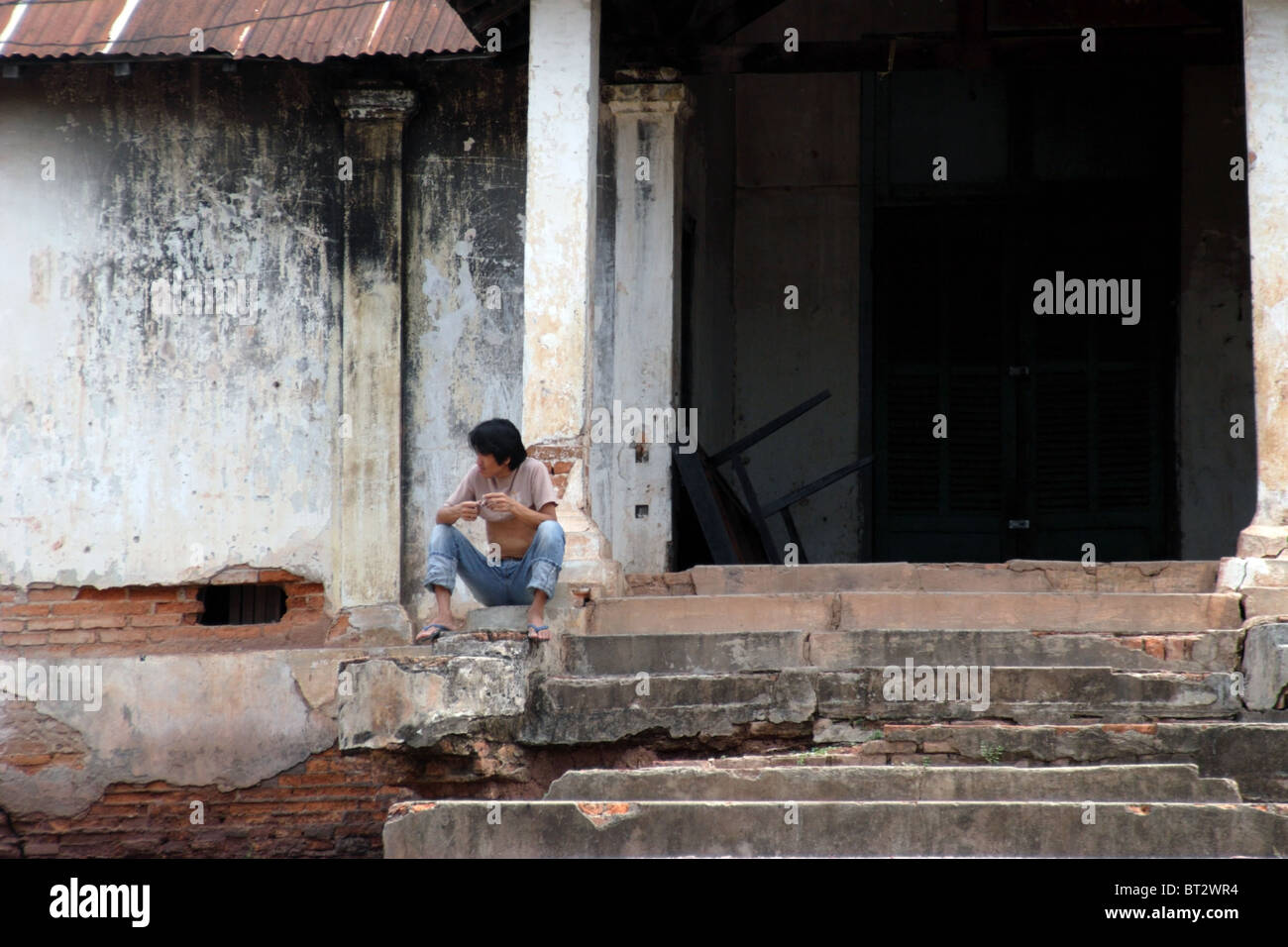 A young man is sitting on the steps of a dilapidated old building in communist Laos. Stock Photo