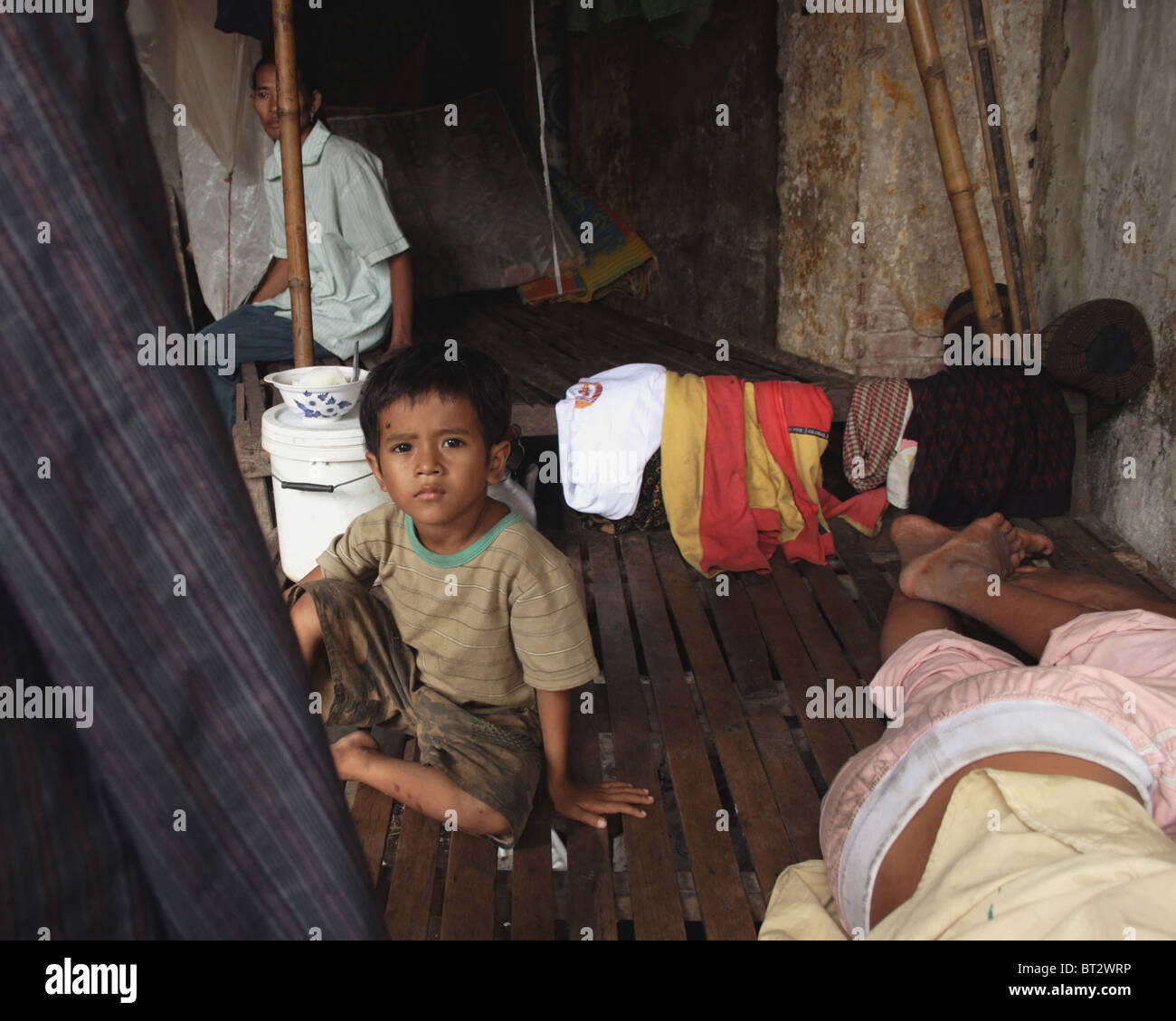 A young child laborer boy living in poverty is sitting on a wooden bed while another boy is sleeping in Kampong Cham, Cambodia. Stock Photo