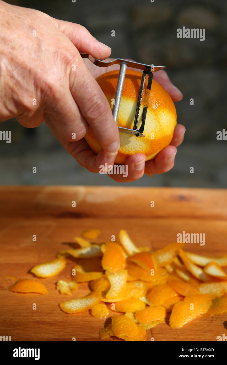 Peeling the rind of an orange by hand with a metal peeler, wooden chopping board Stock Photo