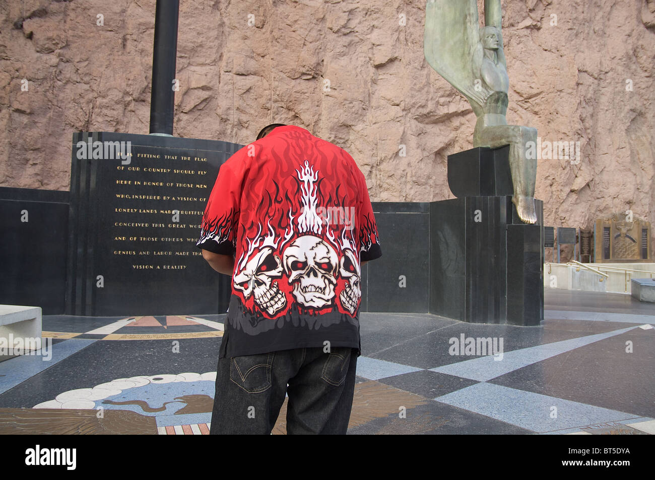 A man in a shirt with flaming skulls on the back pauses near the winged statues at the Hoover Dam Stock Photo