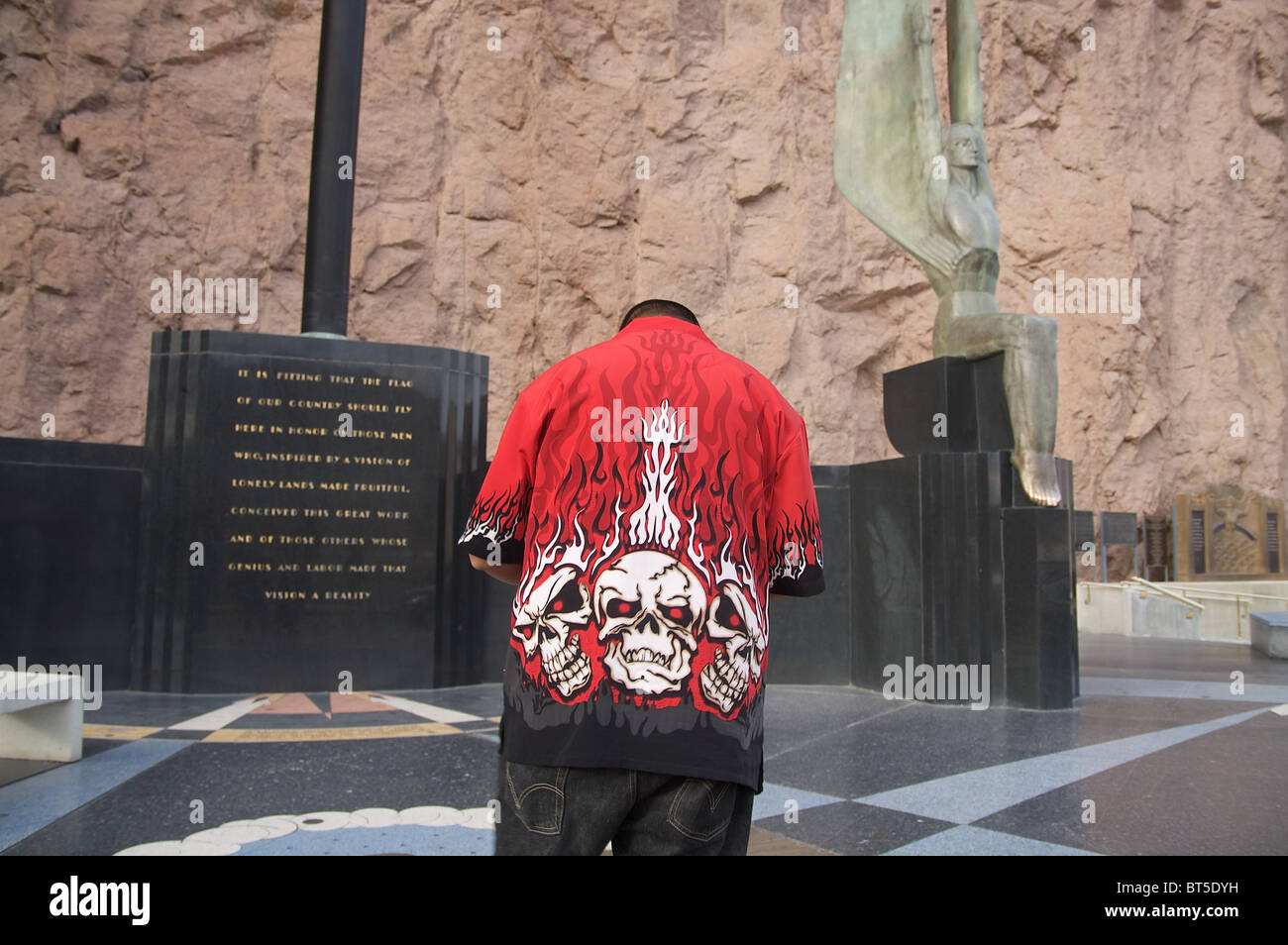 A man in a shirt with flaming skulls on the back pauses near the winged statues at the Hoover Dam Stock Photo