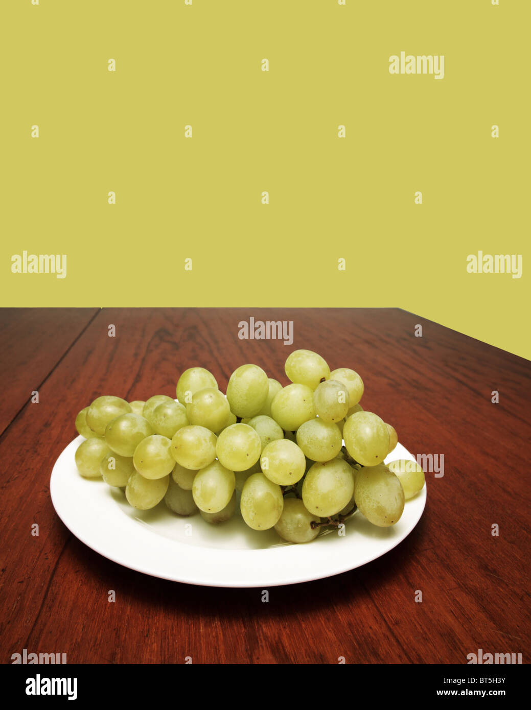 Bunch of white grapes on a white plate on a wooden table with a grape-coloured background Stock Photo