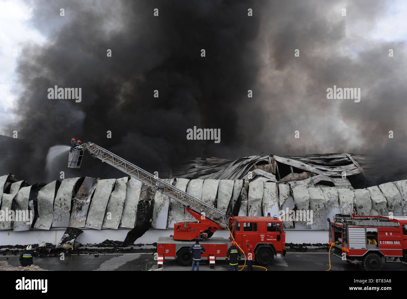 Firemen on a firetruck's extension ladder fighting a massive factory fire amidst thick smoke Stock Photo