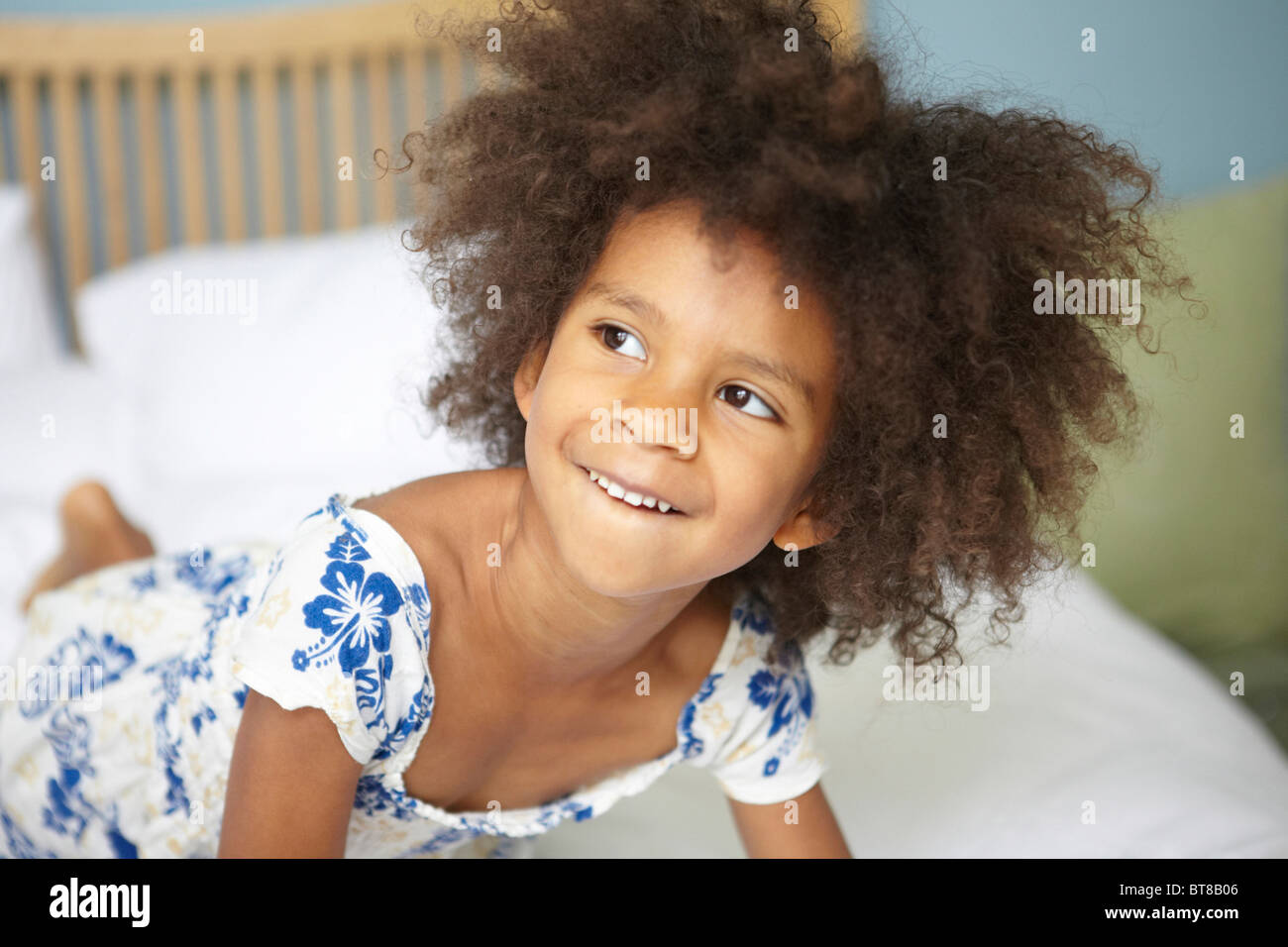 young mixed race girl (6) on bed looking mischievous in blue and white flower dress, leaning forward. Stock Photo