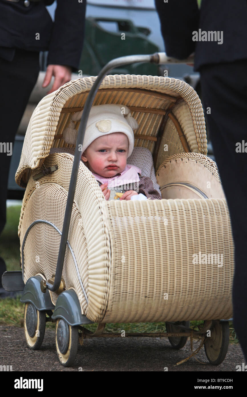 White / Cream Antique Woven Wicker Four Wheel Baby Pram, Folding Handle and Hood and Child looking towards camera Stock Photo