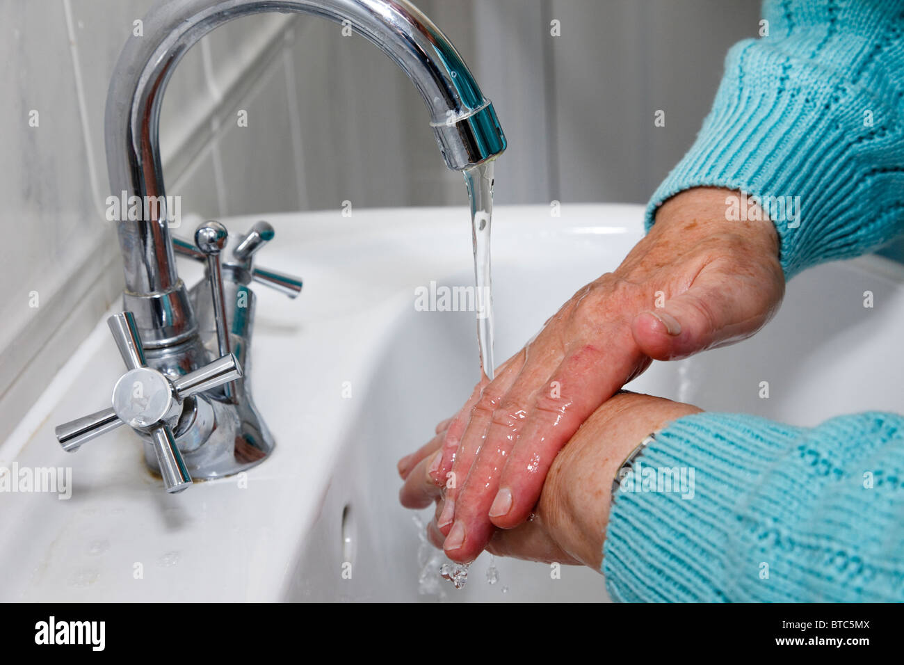 A senior elderly woman cleans by washing hands thoroughly under running tap water in a handbasin to protect against disease germs. England UK Britain Stock Photo