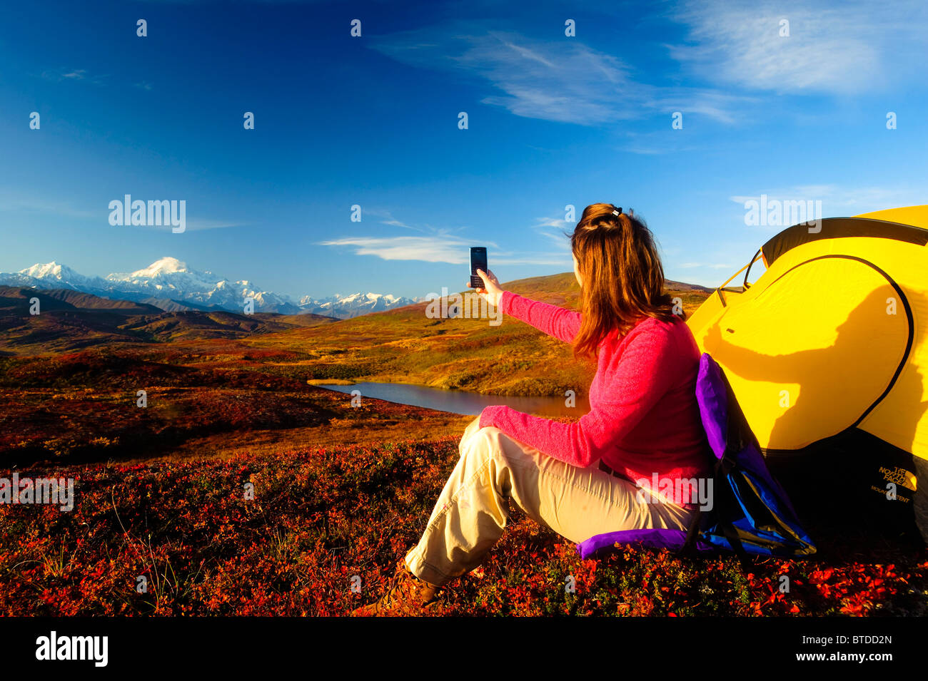 A woman talks on her cell phone and takes a picture of Mt. McKinley near her tent in Peters Hills, Denali State Park,  Alaska Stock Photo