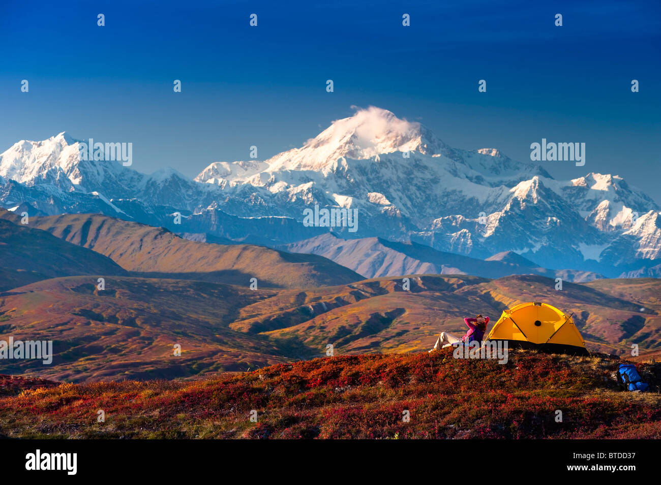 A woman relaxes next to her tent in Peters Hills with a view of Mt. McKinley in the background, Denali State Park,  Alaska, Fall Stock Photo