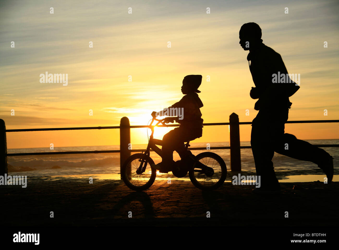 Child riding a bicycle on the Sea point esplanade at sunset with man jogging behind Stock Photo