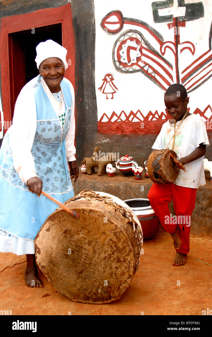 A black Venda woman and small boy beating drums outside a building with mural on the wall and hand made crafts on display Stock Photo
