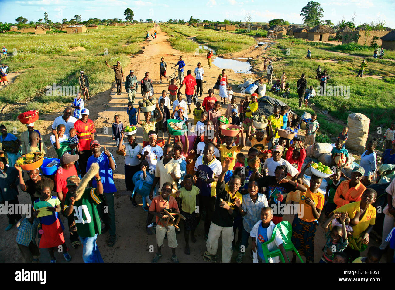 A large group of local villagers, many with bowls of fresh produce gathered in the road of a rural village Stock Photo