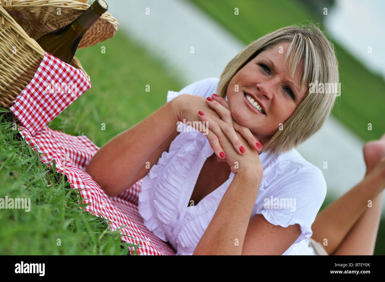 Young blond woman on picnic with wine, basket, gingham blanket and napkin. Stock Photo
