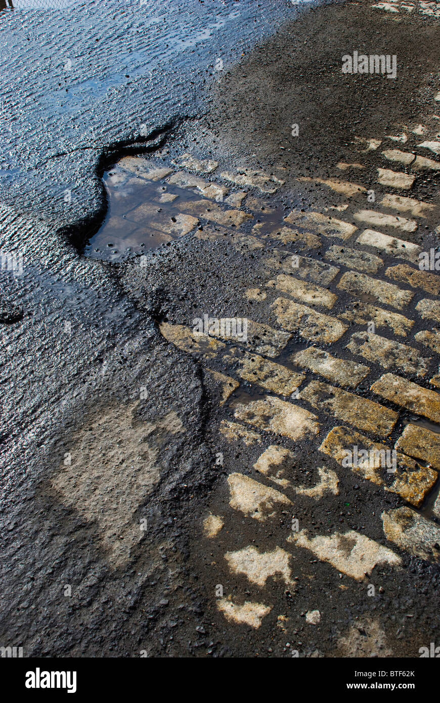 Exposed old stone foundation road beneath roughed macadam awaits resurfacing after a rain. Stock Photo