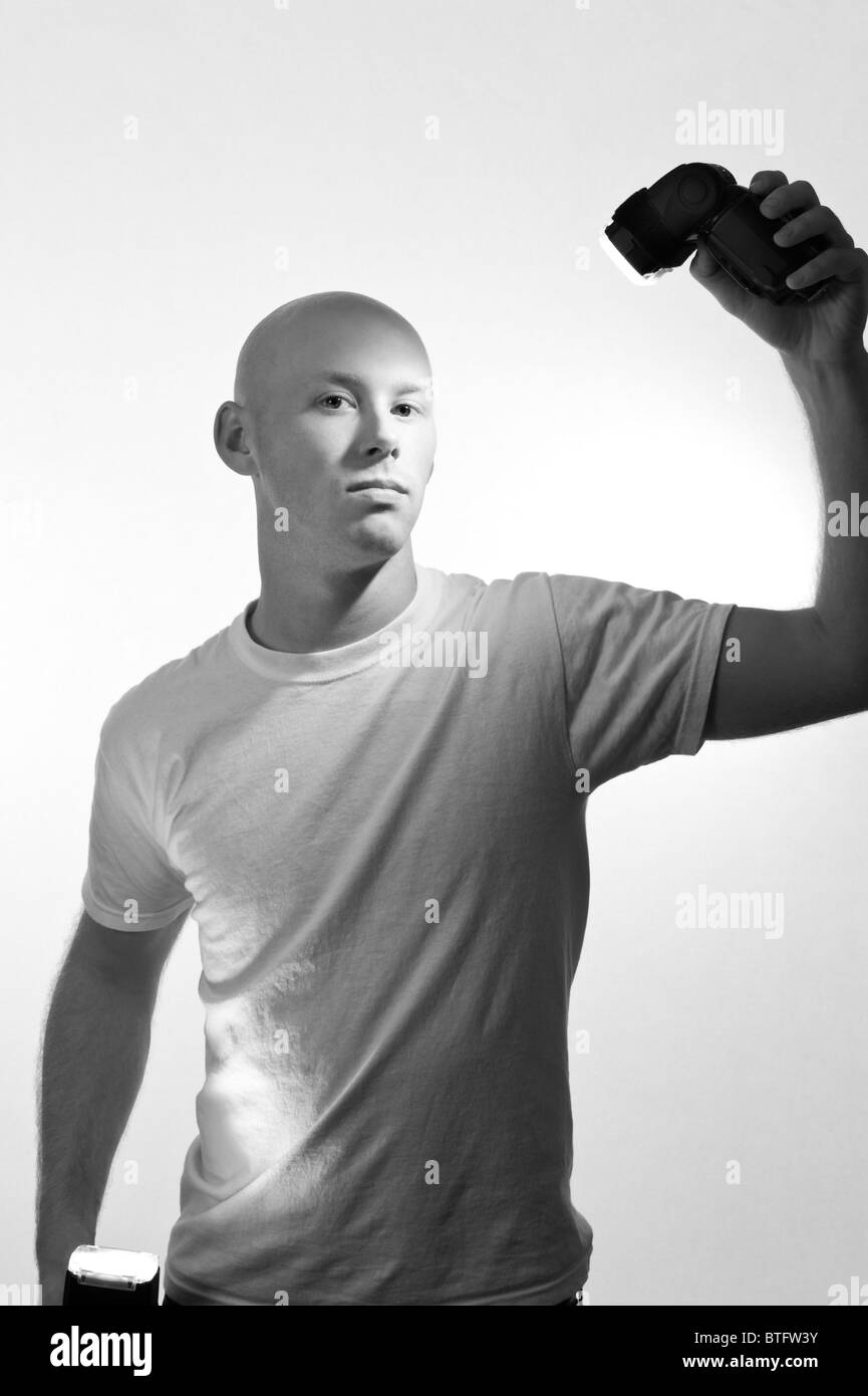 A young bald man in a white tee shirt and white background holding and being lit by two camera flashes. Stock Photo