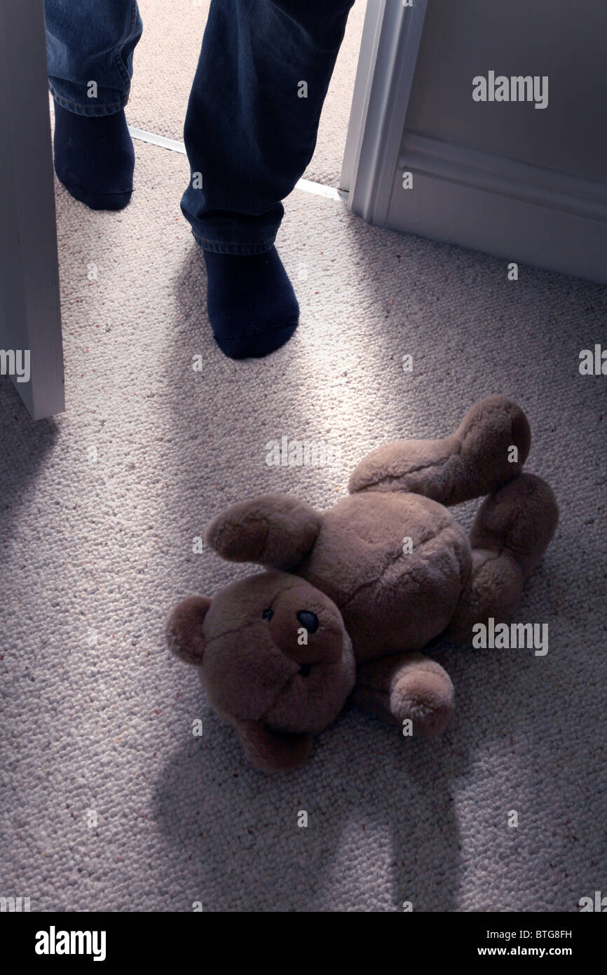 Child's teddy bear lying on the floor of a carpeted room as a man's feet step in through an open door Stock Photo