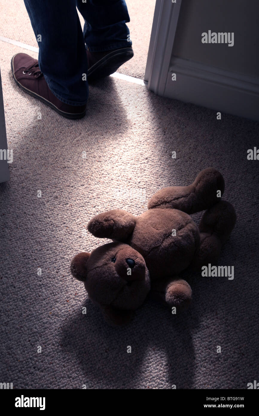 Child's teddy bear lying on the floor of a carpeted room as a man's feet step in through an open door Stock Photo