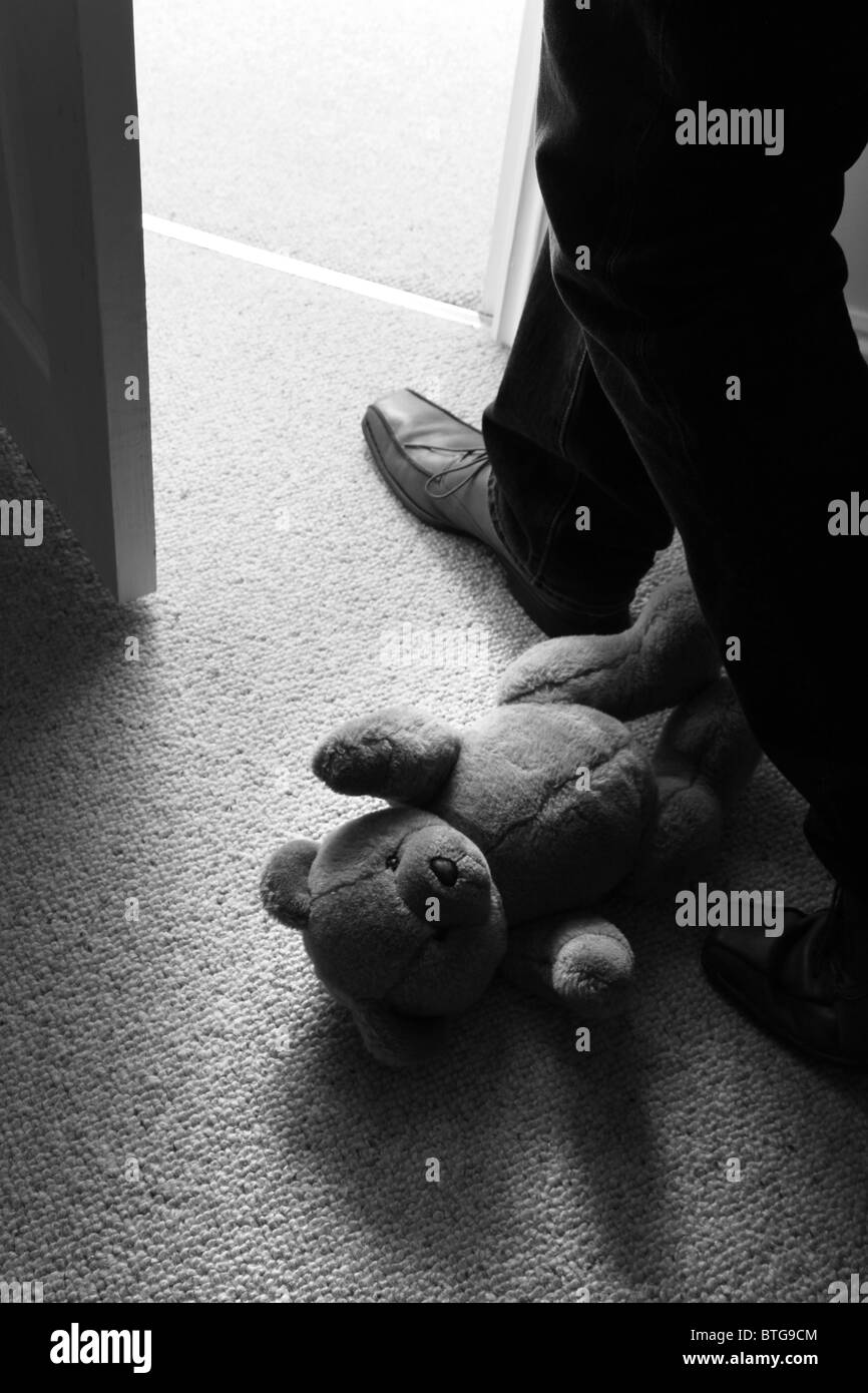 Man (feet and legs only) wearing brown shoes stepping over a teddy bear lying on the floor by an open door. B&W Stock Photo