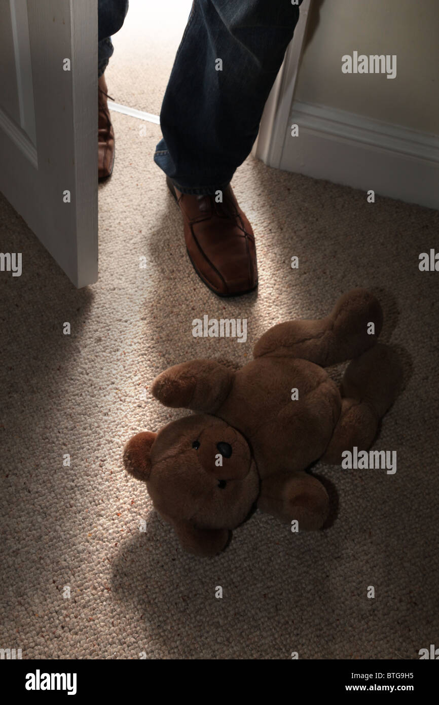 Child's teddy bear lying on the floor of a carpeted room as a man's feet step into an open door Stock Photo