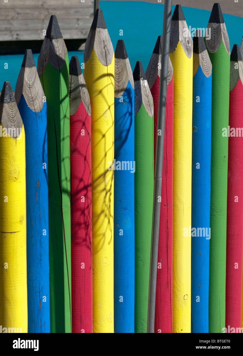 A fence in a school playground in the form of giant coloured pencils with the shadow of a netball net falling across it Stock Photo