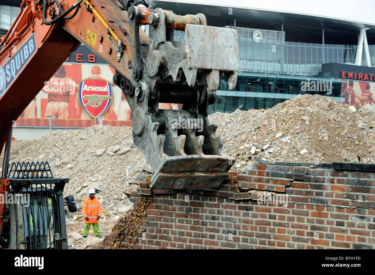 Demolition site showing close up of a hydraulic pulverizer on the arm of an excavator demolishing a wall Arsenal Emirates Stadium in background Stock Photo