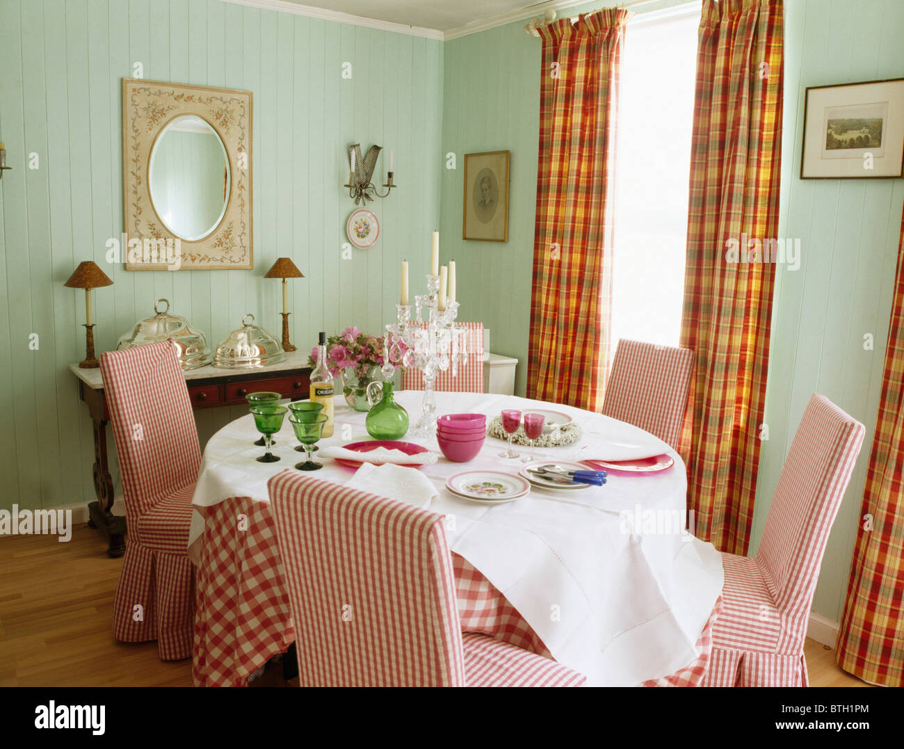 Red checked loose-covers on chairs at table with white linen cloth in pale green dining room with orange-checked curtains Stock Photo