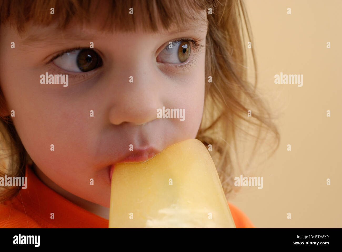 Three year old girl eating an orange ice lolly. Stock Photo