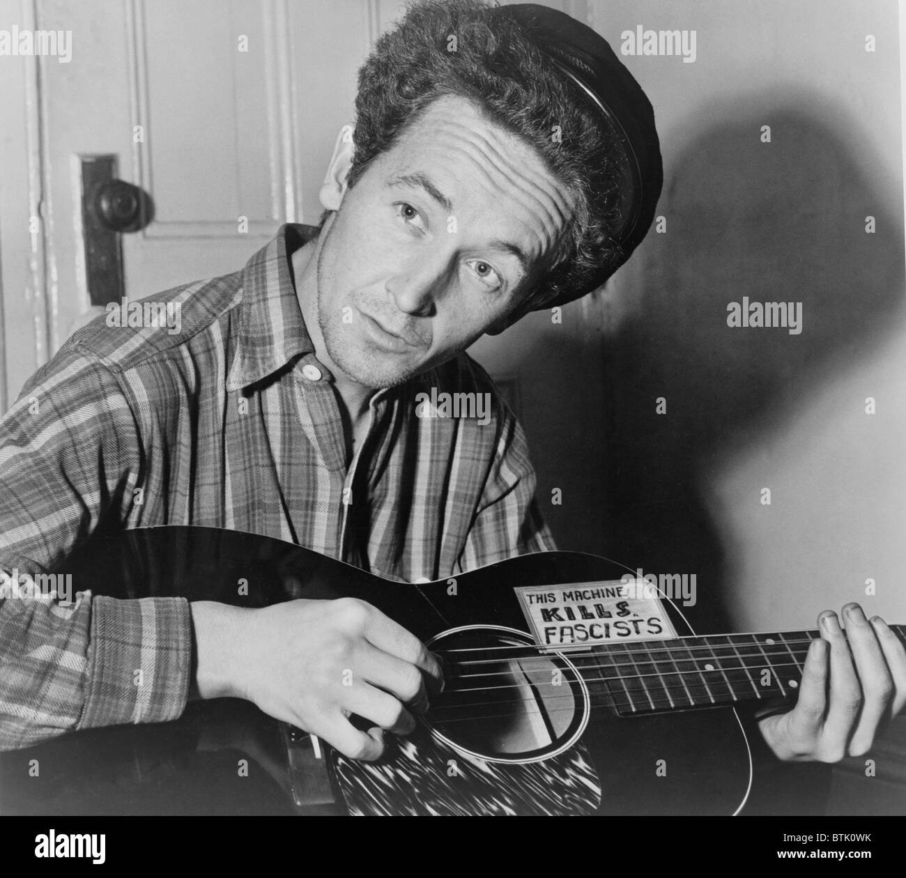 Woody Guthrie (1912-1967), folk singer playing a guitar that has a sticker attached reading: This Machine Kills Fascists. He wrote over 1,000 songs, many still well known, such as THIS LAND IS YOUR LAND. 1943. Stock Photo