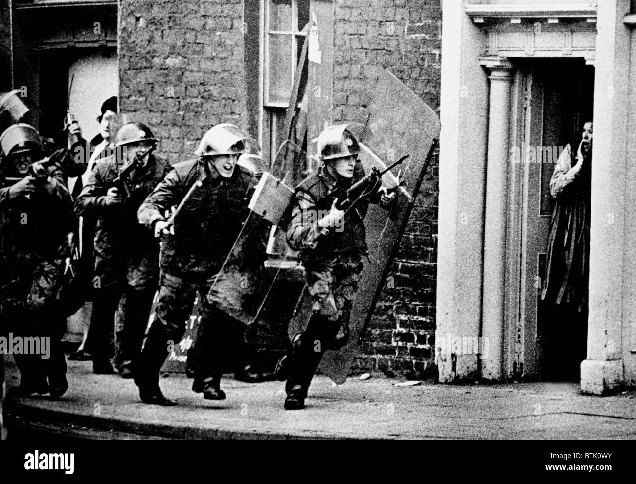 British soldiers chase a sniper down the street in Northern Ireland while a British housewife stands shocked in a doorway Stock Photo
