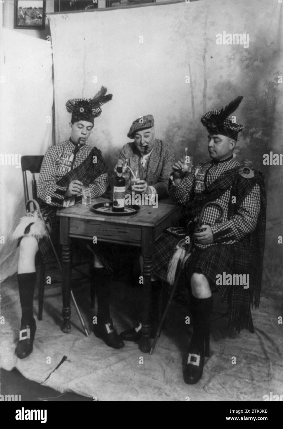 Two men wearing kilts and man smoking cigar, seated at small table drinking Sandy MacDonald scotch, photograph, 1913 Stock Photo