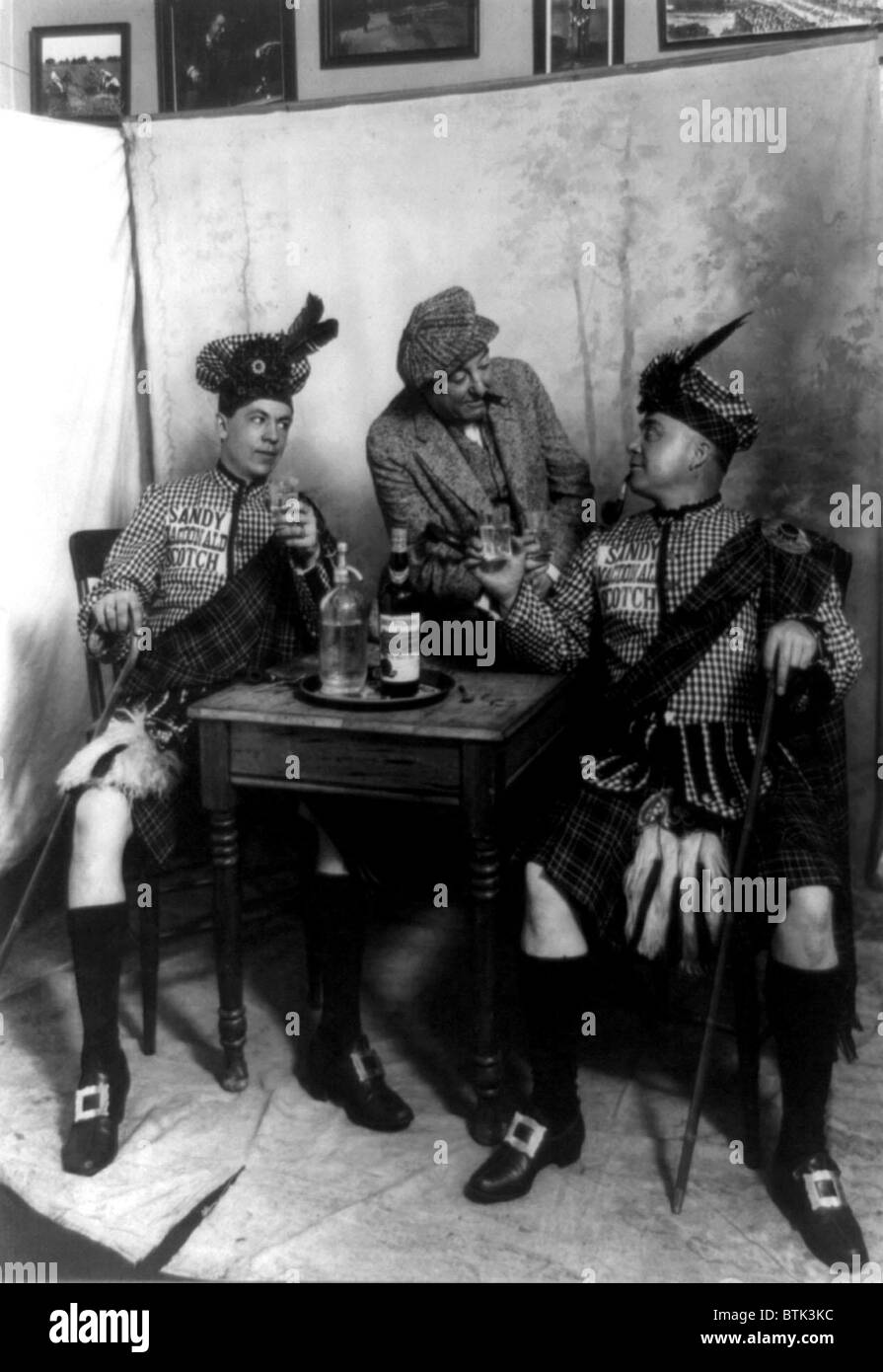 Two men wearing kilts seated at small table, drinking Sandy MacDonald scotch, with man standing and leaning against the table, photograph, 1913 Stock Photo