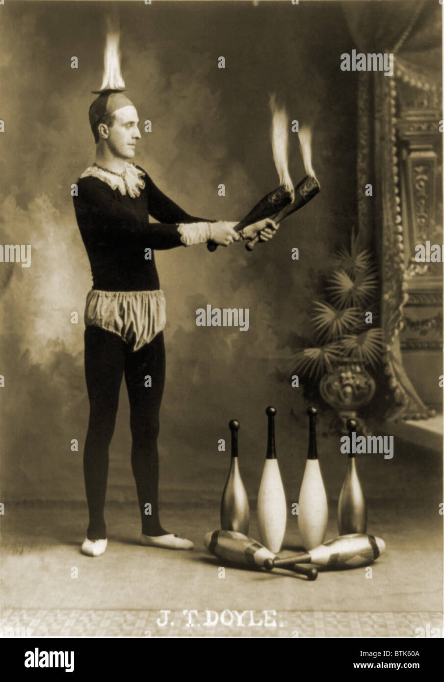 Juggler in jester costume holding two burning clubs while a fire burns on his head.  1902. Stock Photo
