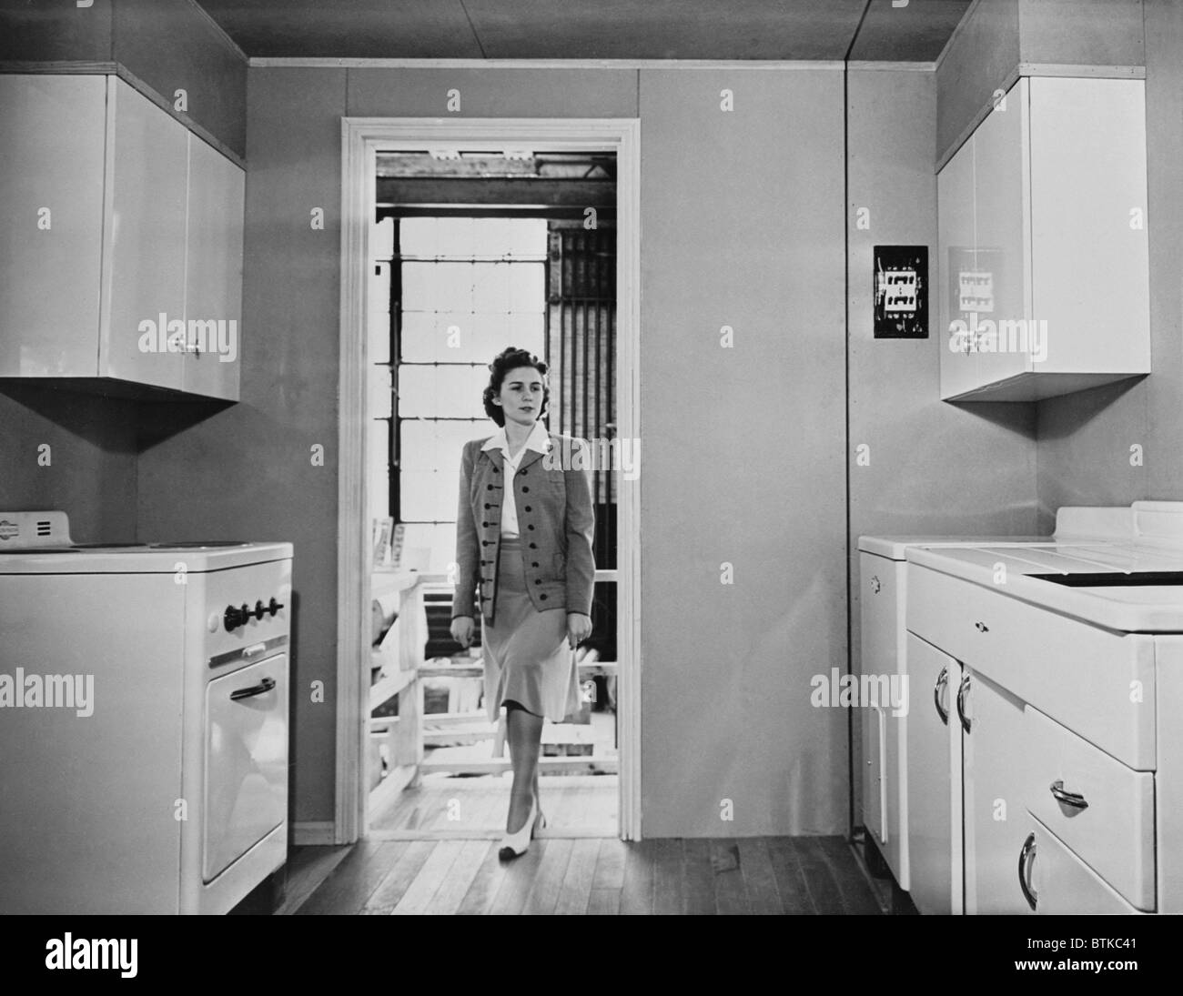 Women enters a modern 1940s kitchen with white enameled metal cabinets.  The housing was built by the government for workers on Stock Photo