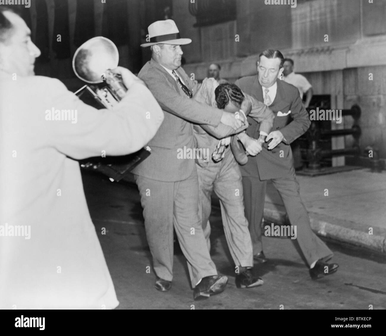 Two policemen forcibly escorting Vincent 'Jimmy Blue Eyes' Ado (1904-2001), after appearing in lineup; a news photographer (left) foreground captures the scene. August 1947. Stock Photo