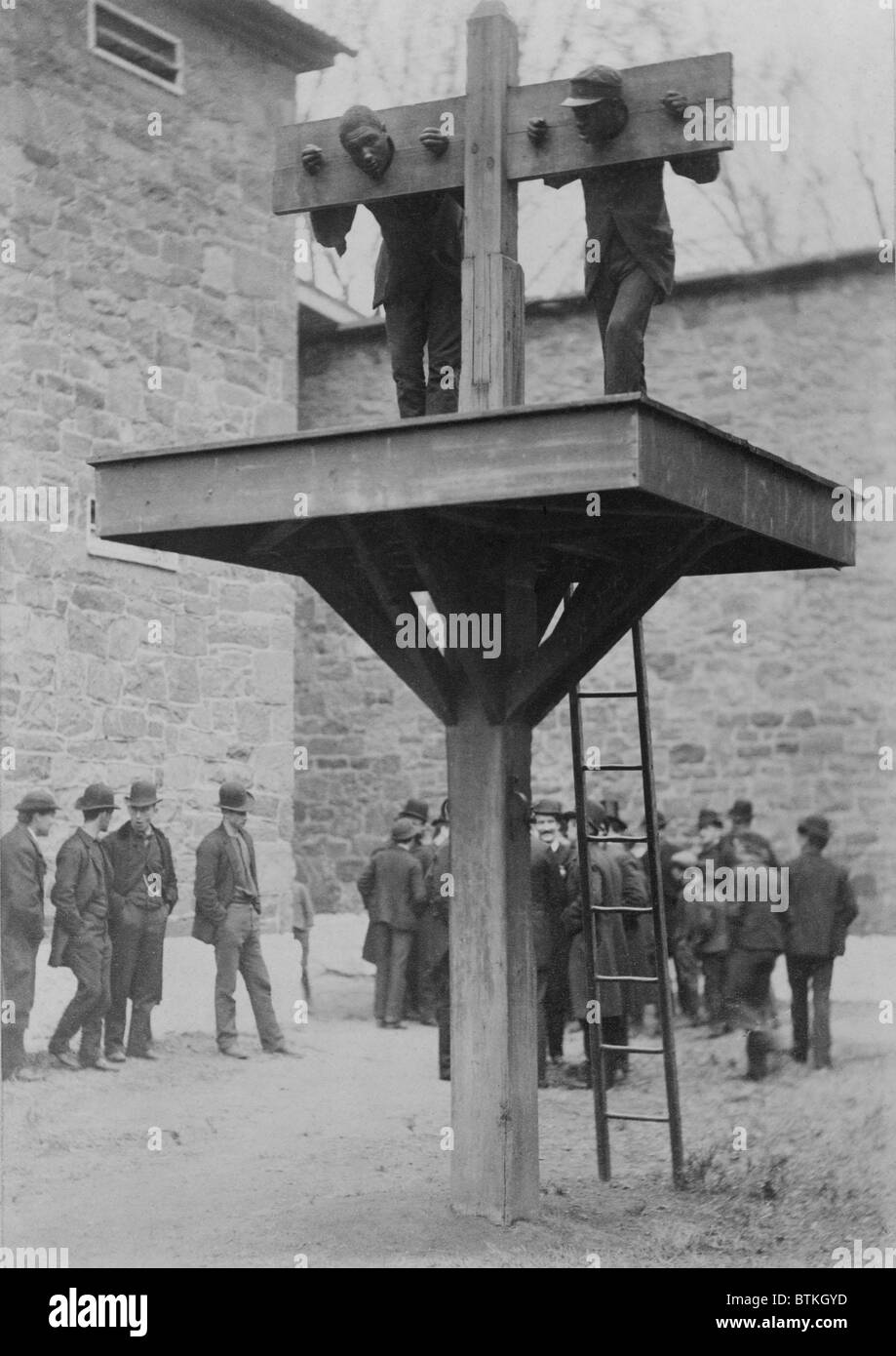 Two African American men confined in a Delaware pillory above a whipping post. Spectators to the punishment loiter in the background. 1889. Stock Photo
