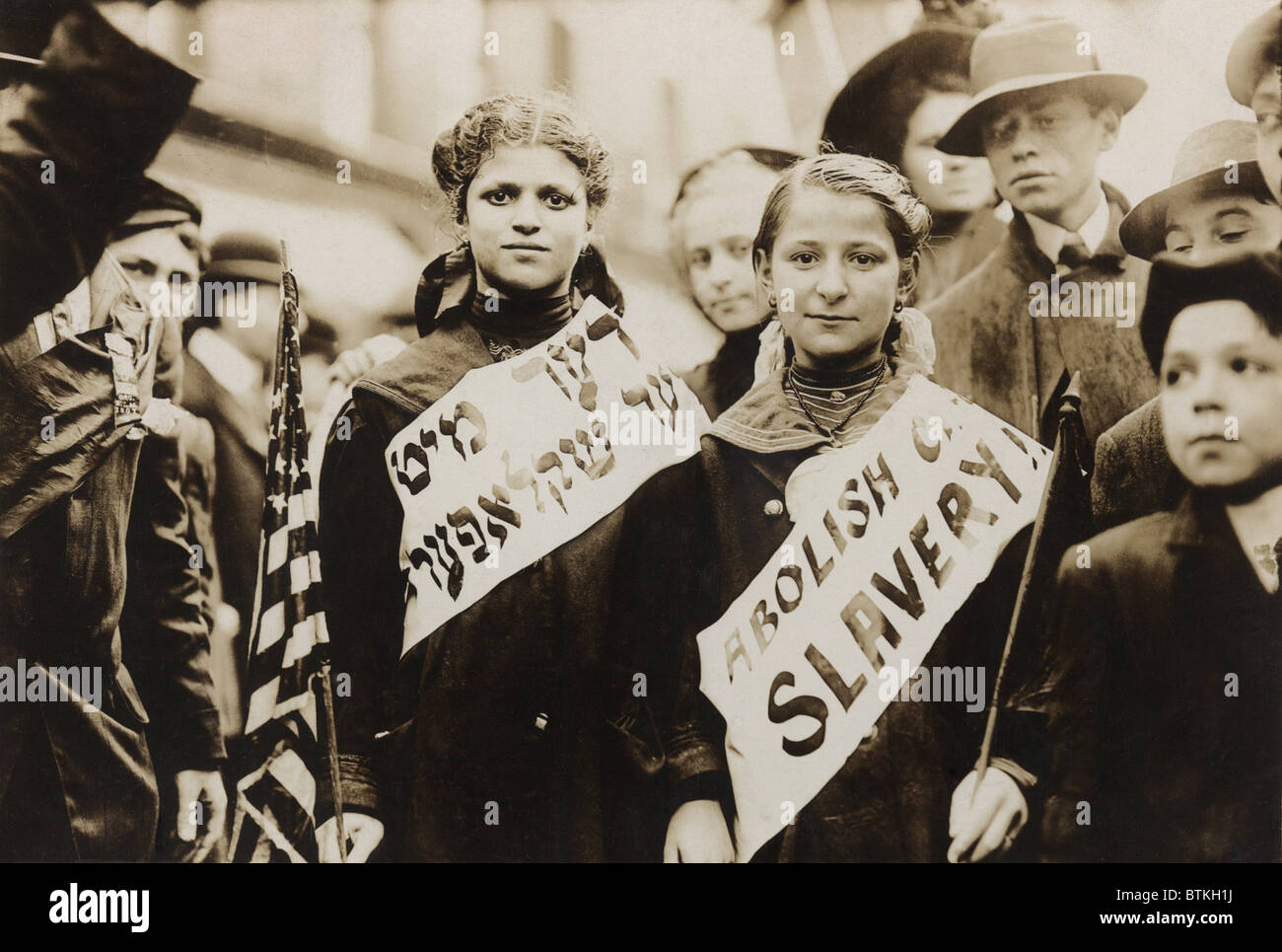New York City May Day celebration of the international labor day. Two girls in the parade combined carrying an American flag, with wearing banners with the slogan ABOLISH CHILD SLAVERY!! In English and Yiddish. 1909. Stock Photo