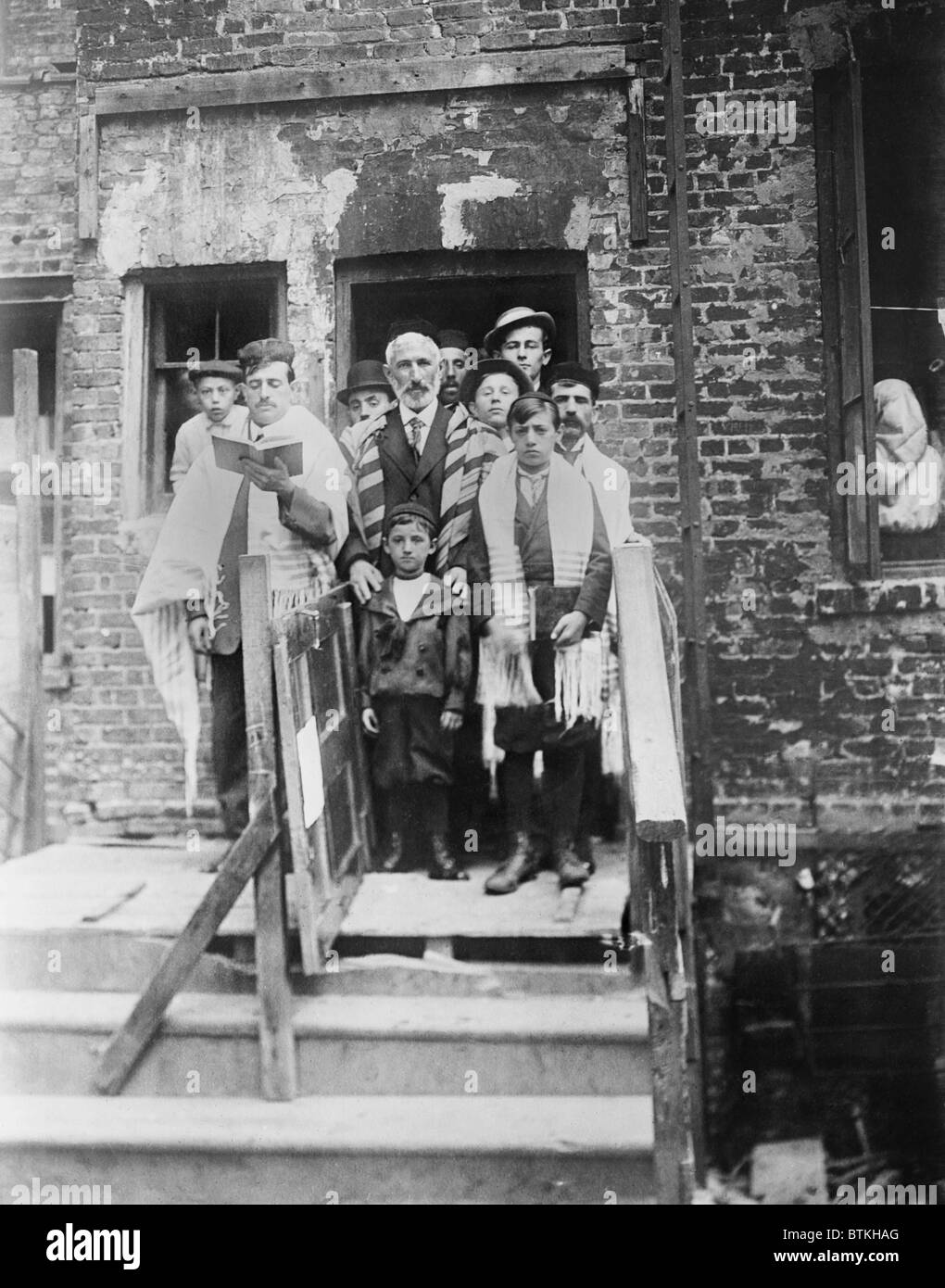 Jewish men and boys in prayer shawls standing in front of a dilapidated tenement synagogue on Yom Kippur. New York City. Ca.1910. Stock Photo