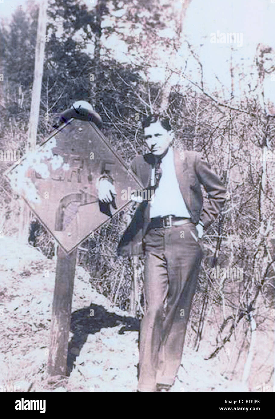 Clyde Barrow (1909-1934) dressed in a suit and standing with his hand place through a hold on a road sign. Stock Photo