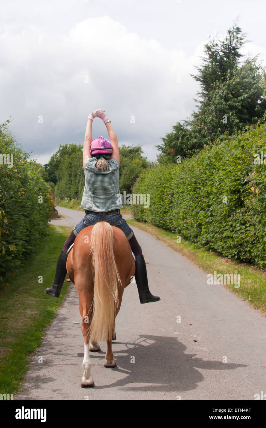 Young woman stretching her arms high in the air as she rides her horse along country lanes in rural Shropshire Stock Photo