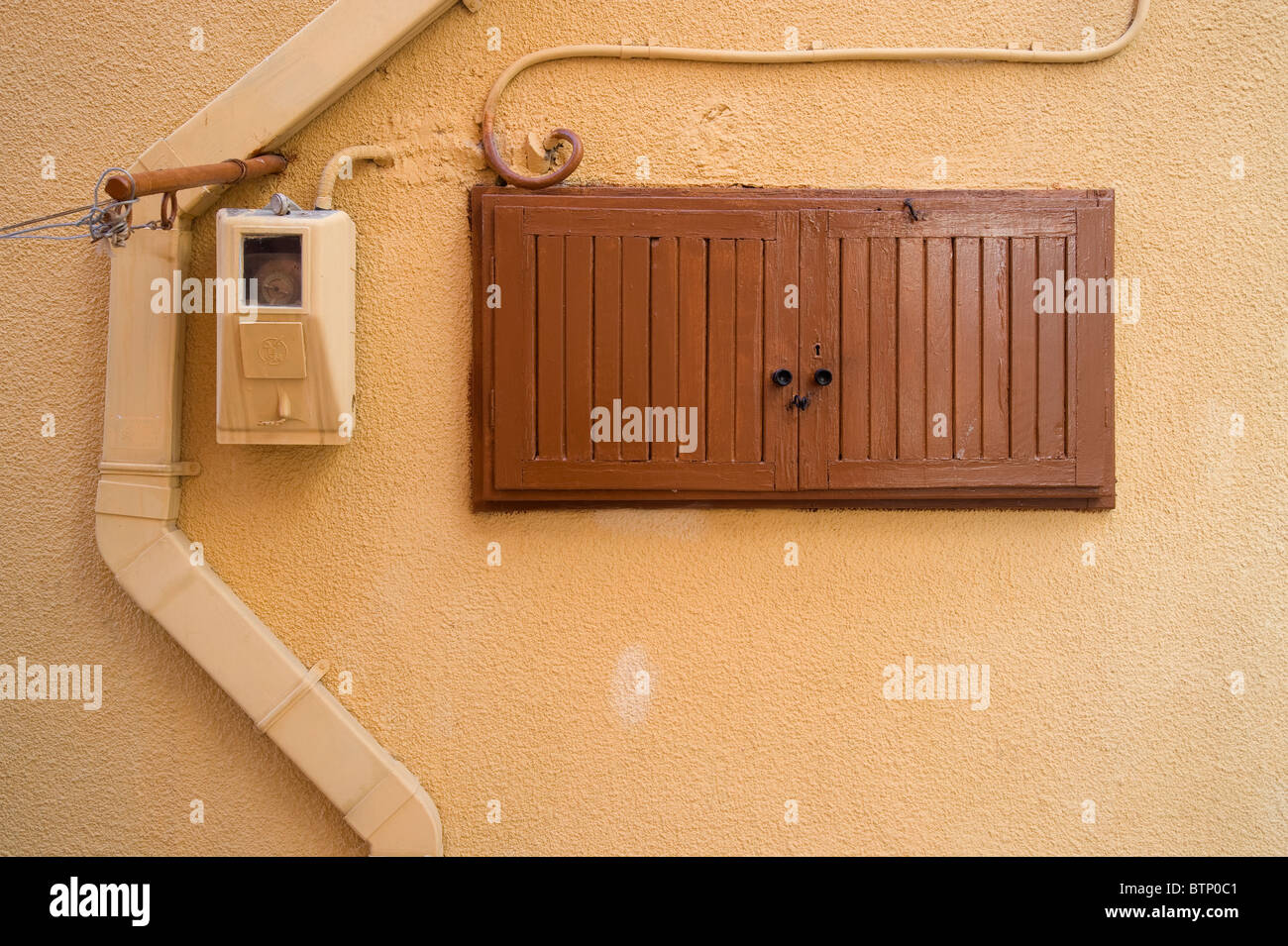 Ocre color adobe wall with electrical meter and closet with brown wooden shutters, in Ermoupolis, on the Greek island of Syros. Stock Photo