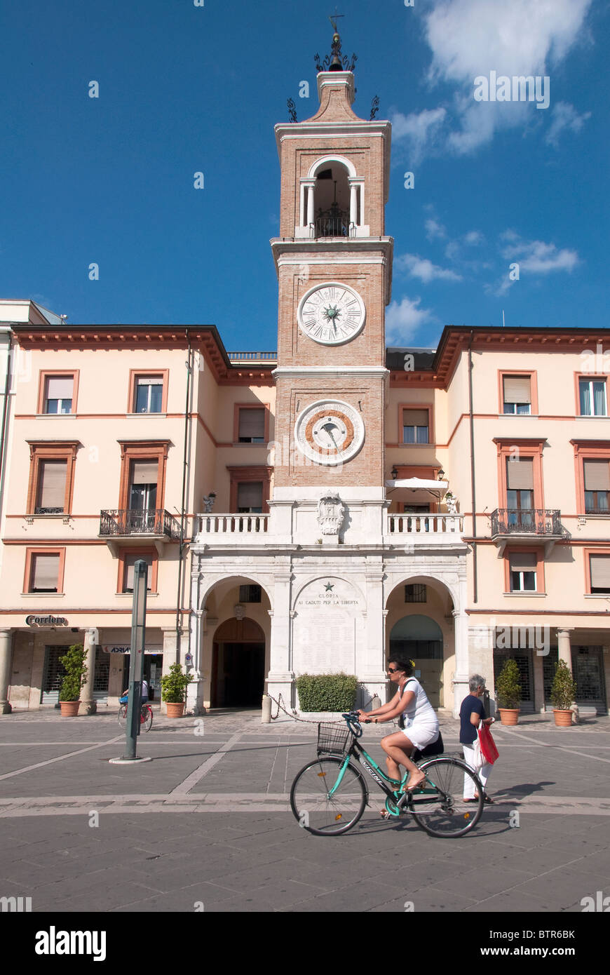 Woman cycling past Clock tower telling the time, date and Zodiac sign in Piazza Cavour, Rimini, Emilia Romagna, Italy Stock Photo