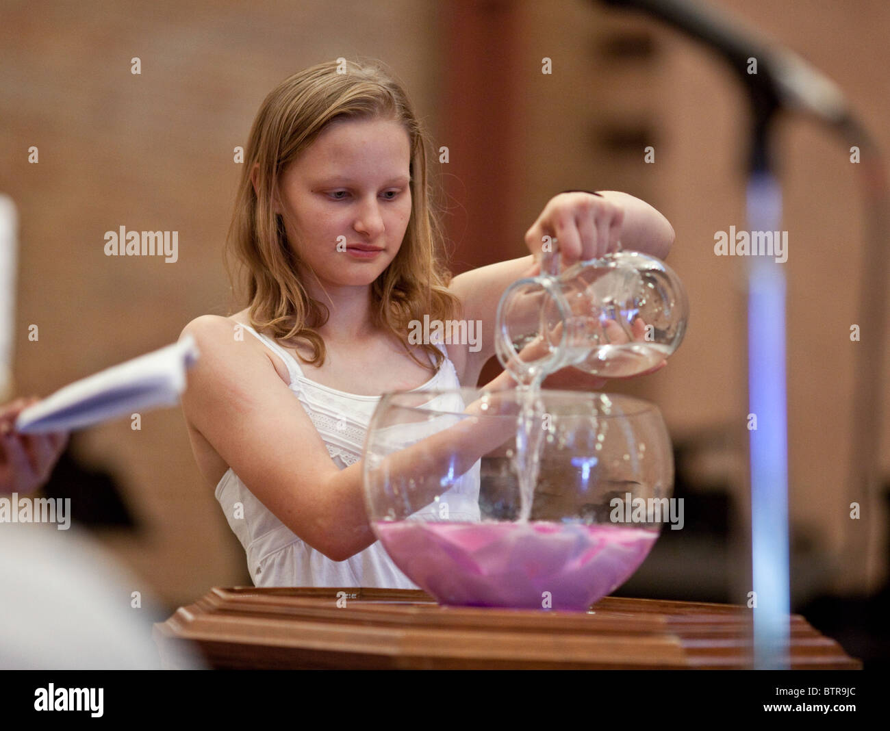 Anglo female teen pours baptismal water into bowl with pink slips of paper with wishes written on them during church service Stock Photo