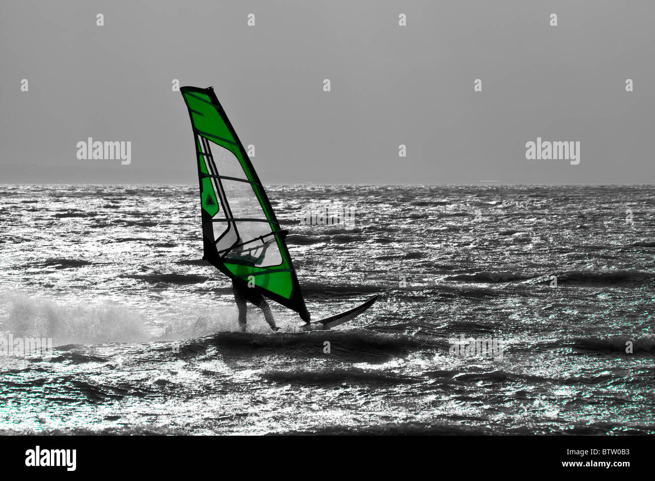 Lone windsurfer silhouetted against grey sky with color of sail enhanced. Stock Photo