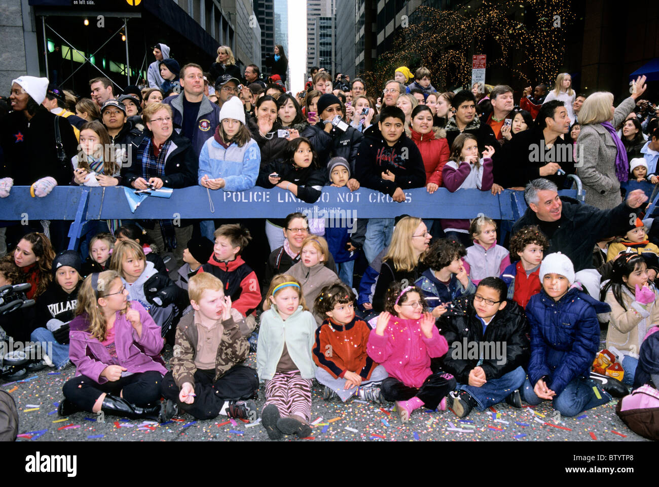 New York City People sitting and standing on the street watching Macy's Thanksgiving Day parade behind a police barrier. Stock Photo