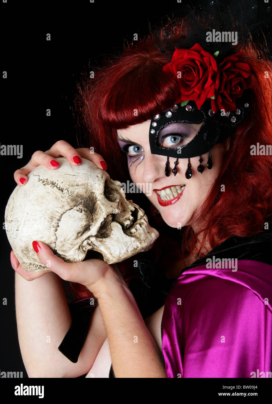 Young Red Headed Woman Wearing a Purple Cape and Sequined Mask Over One Eye and a Red Rose and Holding A Human Skull Stock Photo