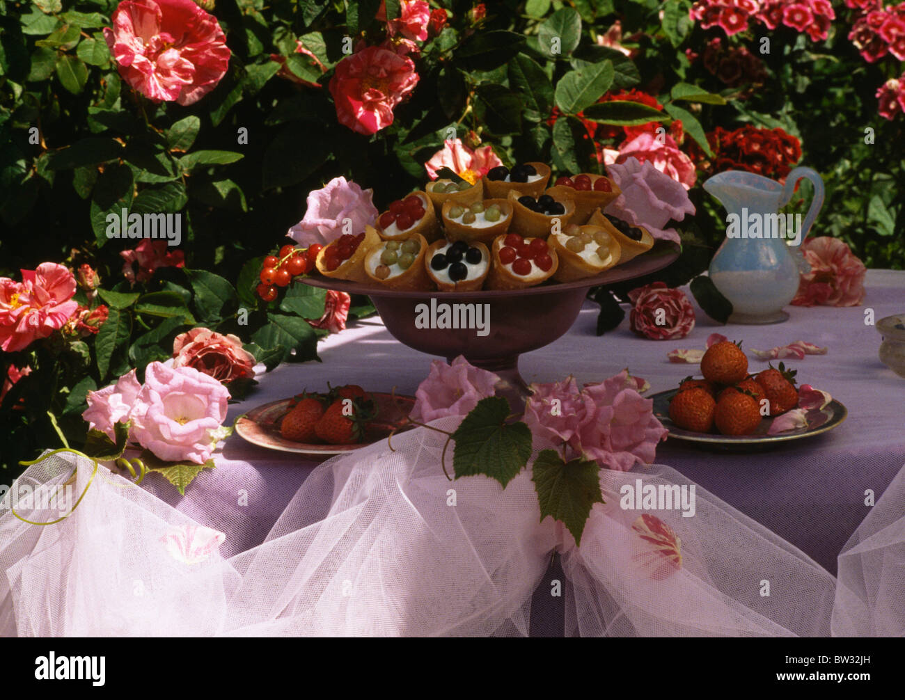 Still-Life of small cakes on pedestal on garden table swathed with white organza and decorated with pink roses Stock Photo