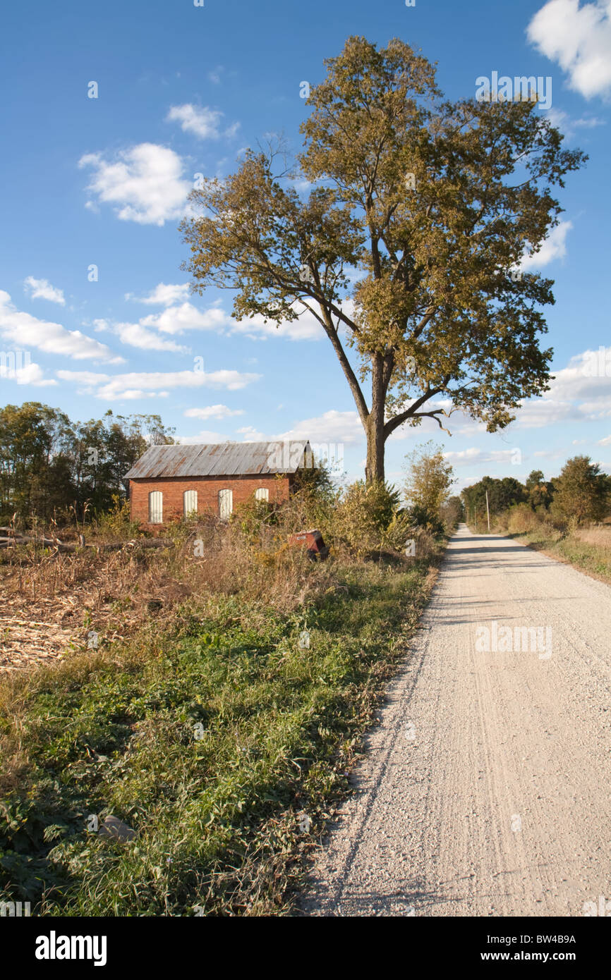 Abandoned on-room schoolhouse on a rural, gravel road in Indiana with a large tree, bright blue sky and clouds vertical Stock Photo
