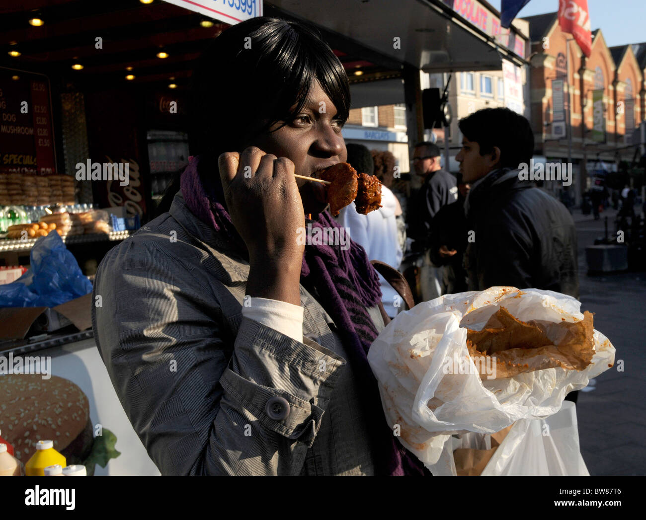 UK AFRO CARIBBEAN WOMAN EATING AT FOOD STALL IN THE STREETS OF BARKING, LONDON Stock Photo